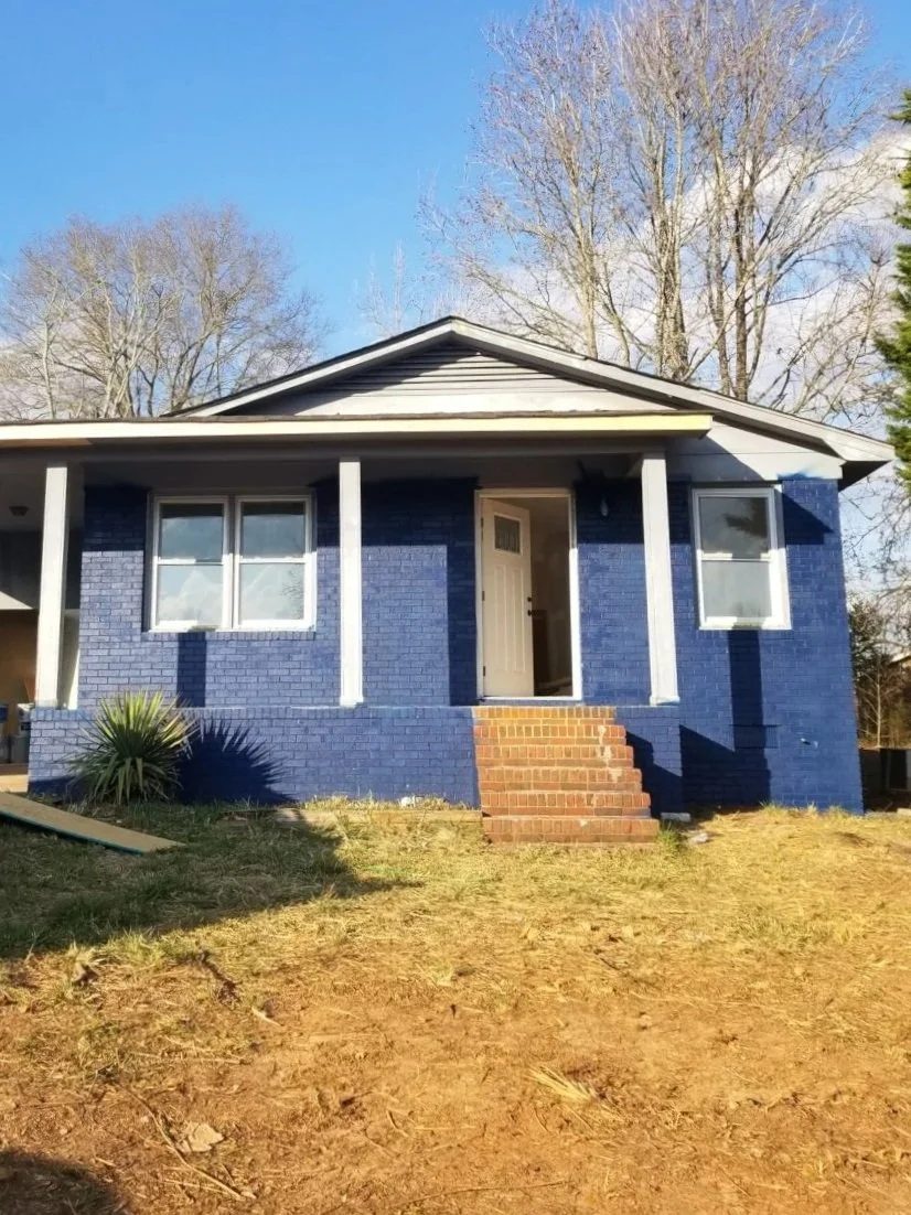 A blue brick house with white trim and a small front porch with three steps leading up to the front door. The house has two windows on either side of the entrance and is situated in a yard with some grass and trees in the background.