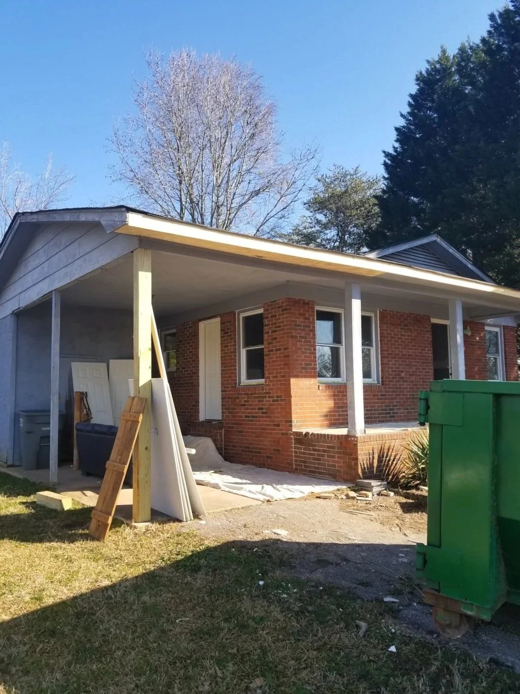 House undergoing renovation with construction materials and tools in front, green trash dumpster nearby, clear blue sky, trees in the background.