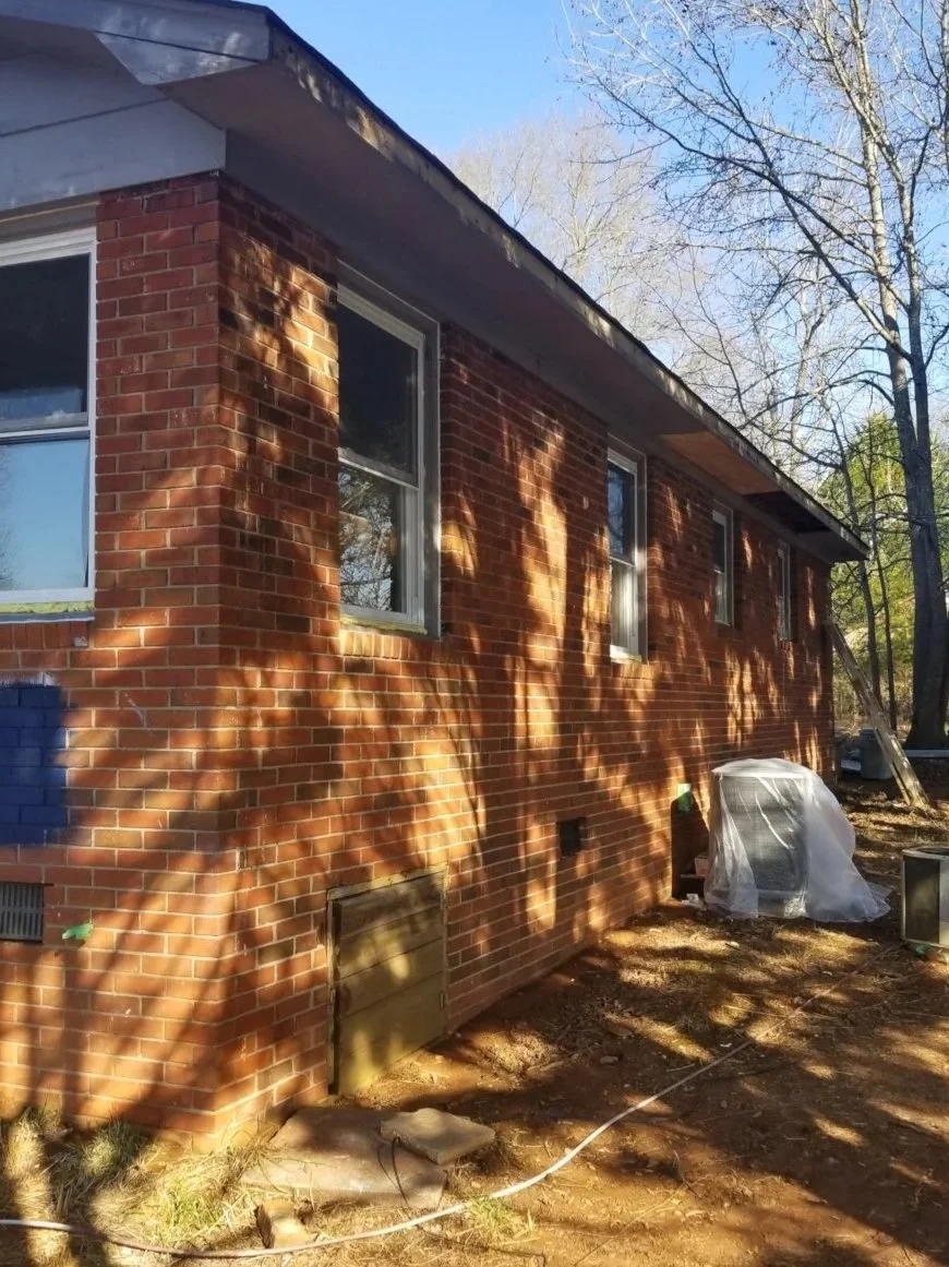 Side of a brick house with three windows, showing sunlight and shadows on the wall, and some construction or renovation materials outside.