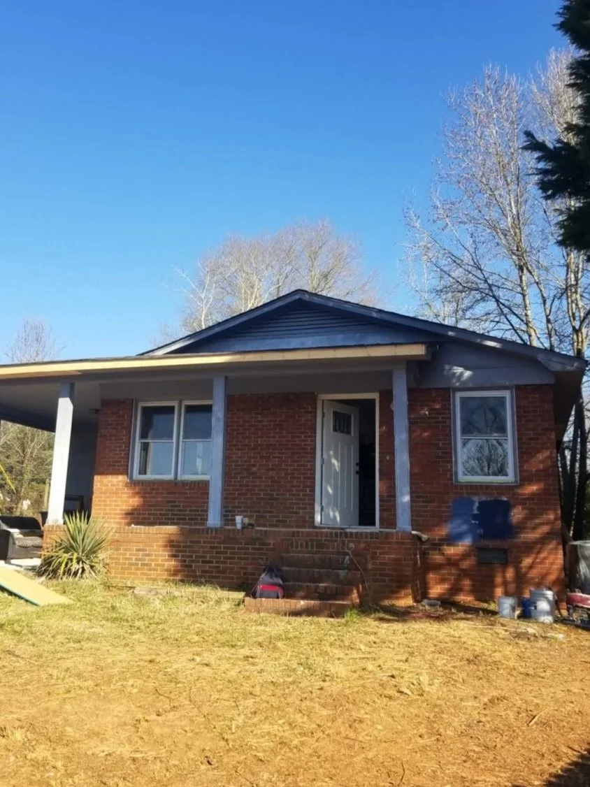 A brick house with a covered front porch, two front windows, a white front door, and a yard with some steps leading to the entrance, on a clear day.