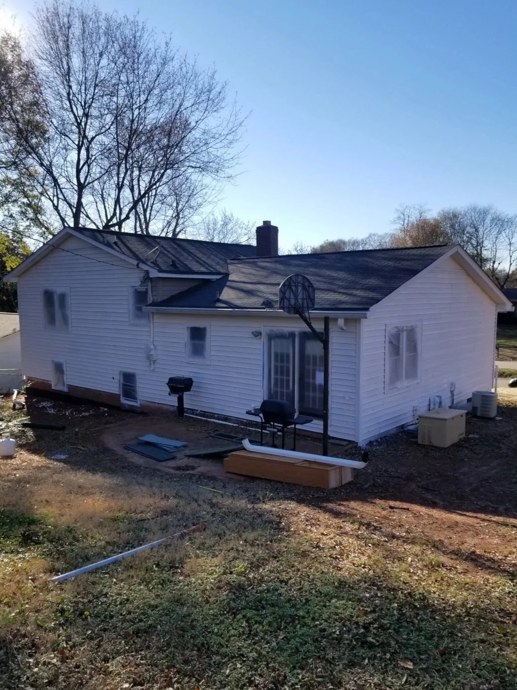 Backyard of a white house with vinyl siding, a sliding glass door, and multiple windows covered in plastic during construction. There is a basketball hoop, a grill, and various construction materials on the ground. Several trees are visible in the ba