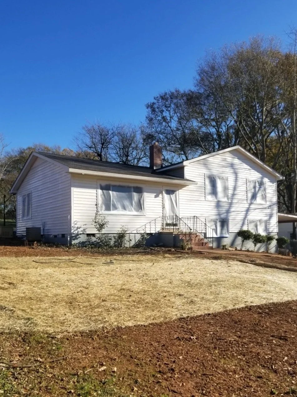A white two-story house with a porch, steps, and black metal railing, surrounded by brown yard soil and trees with bare branches, under a clear blue sky.