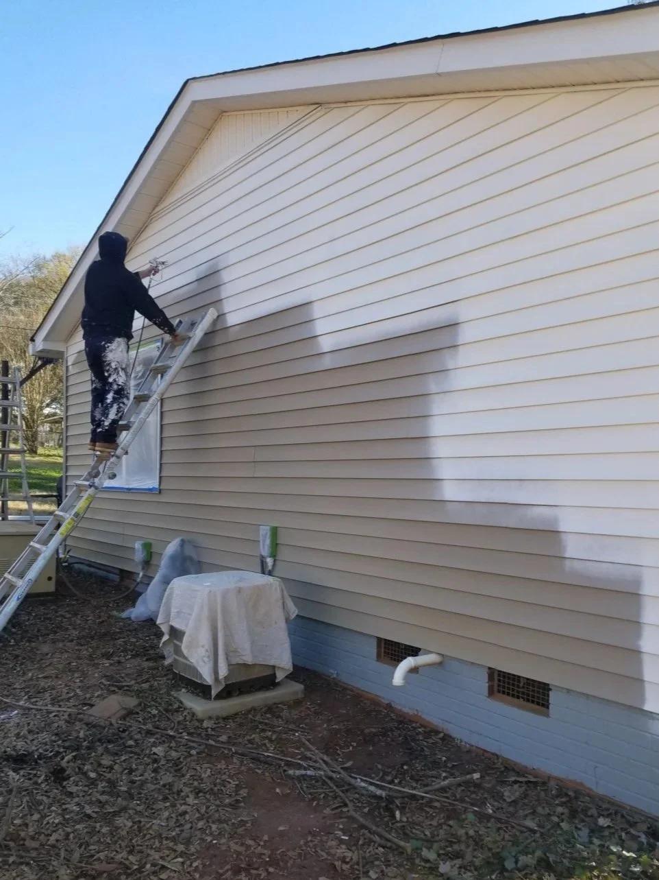 Person on a ladder pressure washing the exterior wall of a house with beige siding.