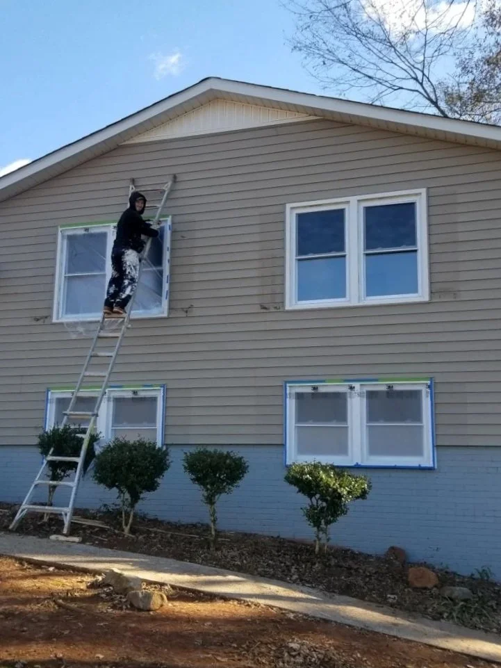 Person on ladder painting or cleaning exterior window of a beige house with tan siding, with multiple windows, bushes in front, and clear blue sky overhead.