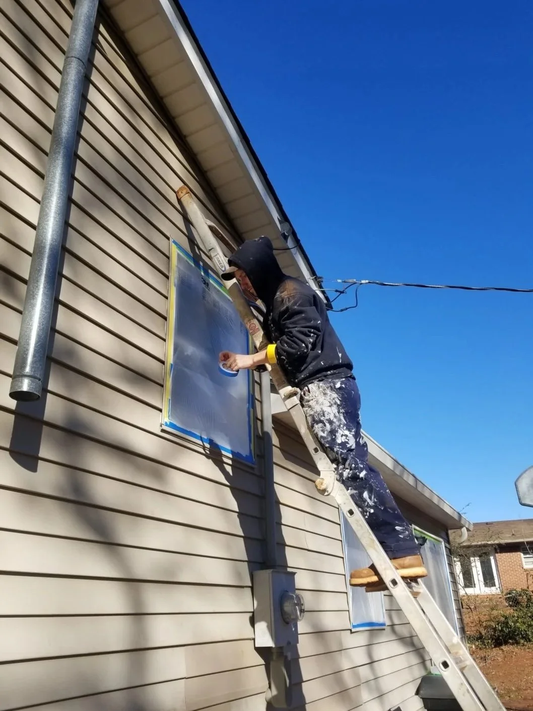 A person on a ladder painting or cleaning a window on the side of a house under a clear blue sky.