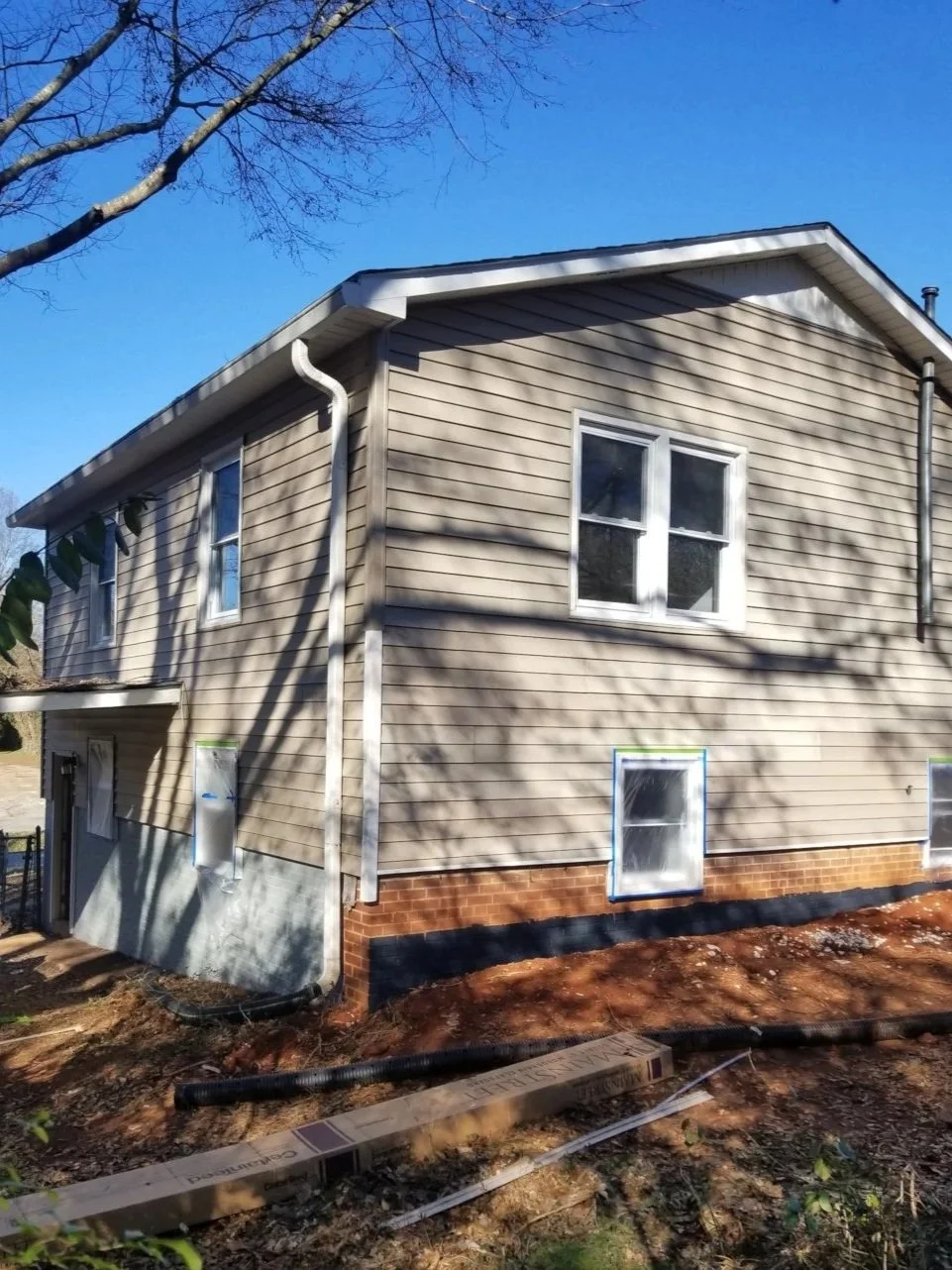 A two-story house with beige siding and a brick foundation, with three visible white-framed windows and a white gutter, surrounded by bare trees and a dirt yard with construction materials on the ground.