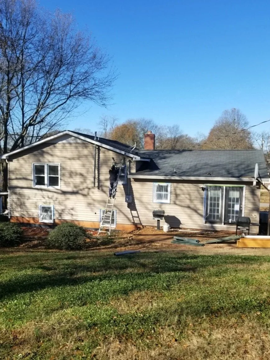 A person on a ladder working on the exterior of a beige house with a black roof, surrounded by trees and a lawn.