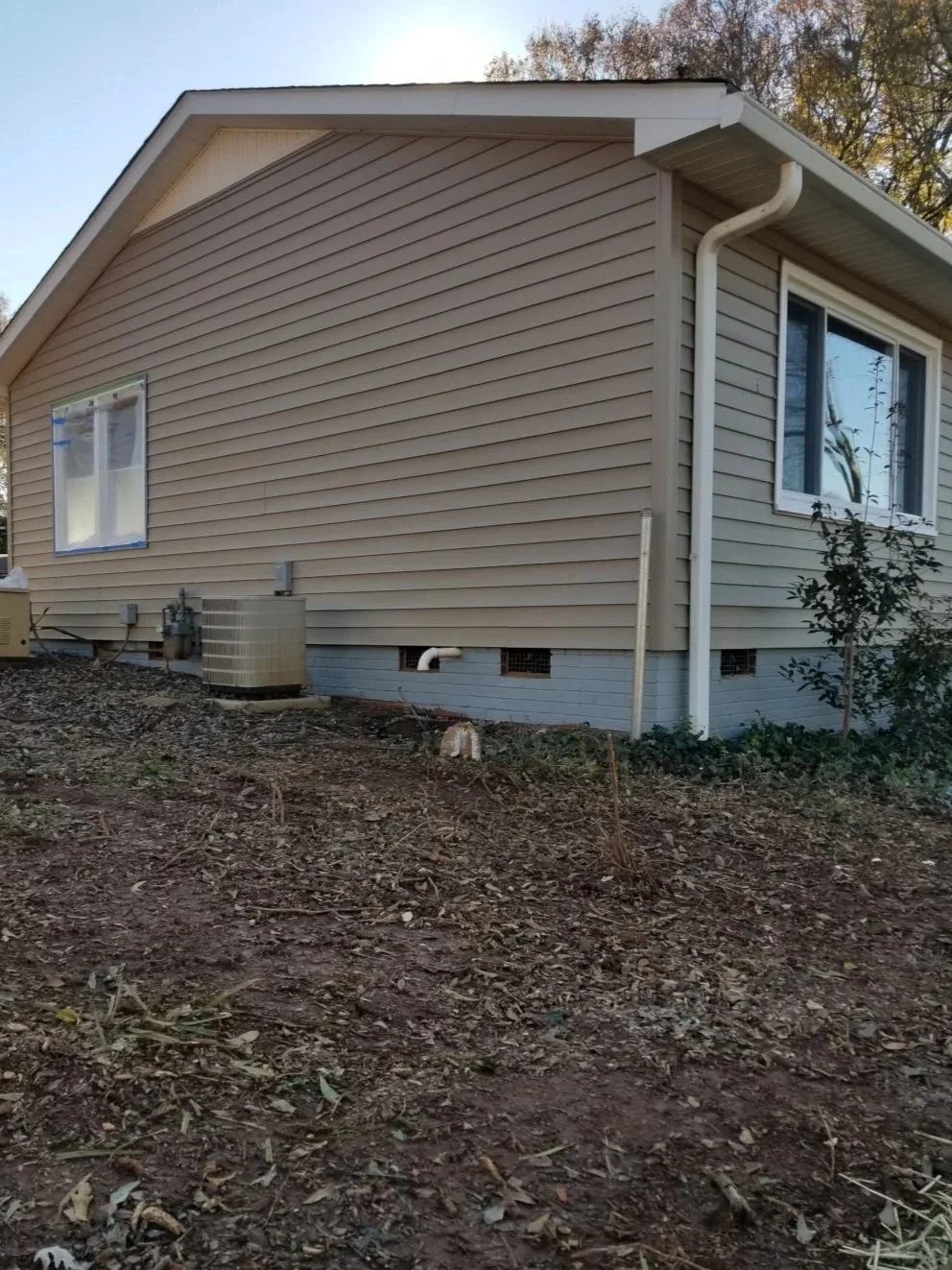 The side of a house with beige siding, two windows, a white rain gutter, and a small patch of ground with dirt and leaves.