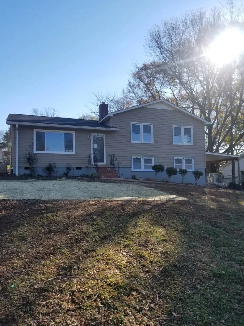 A two-story house with gray siding and white trim, front steps leading to the door, and a lawn with some bushes and trees in the background under a clear sky with bright sunlight.