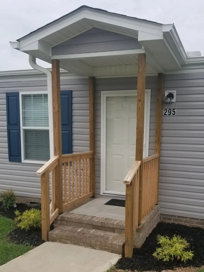 Small front porch with railroad ties, supported by wooden posts, leading to a white door on a gray house with blue shutters.