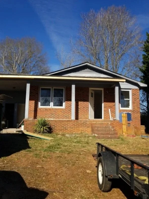 Front view of a brick house with a porch, steps, and two columns, under a blue sky with tall leafless trees in the background.