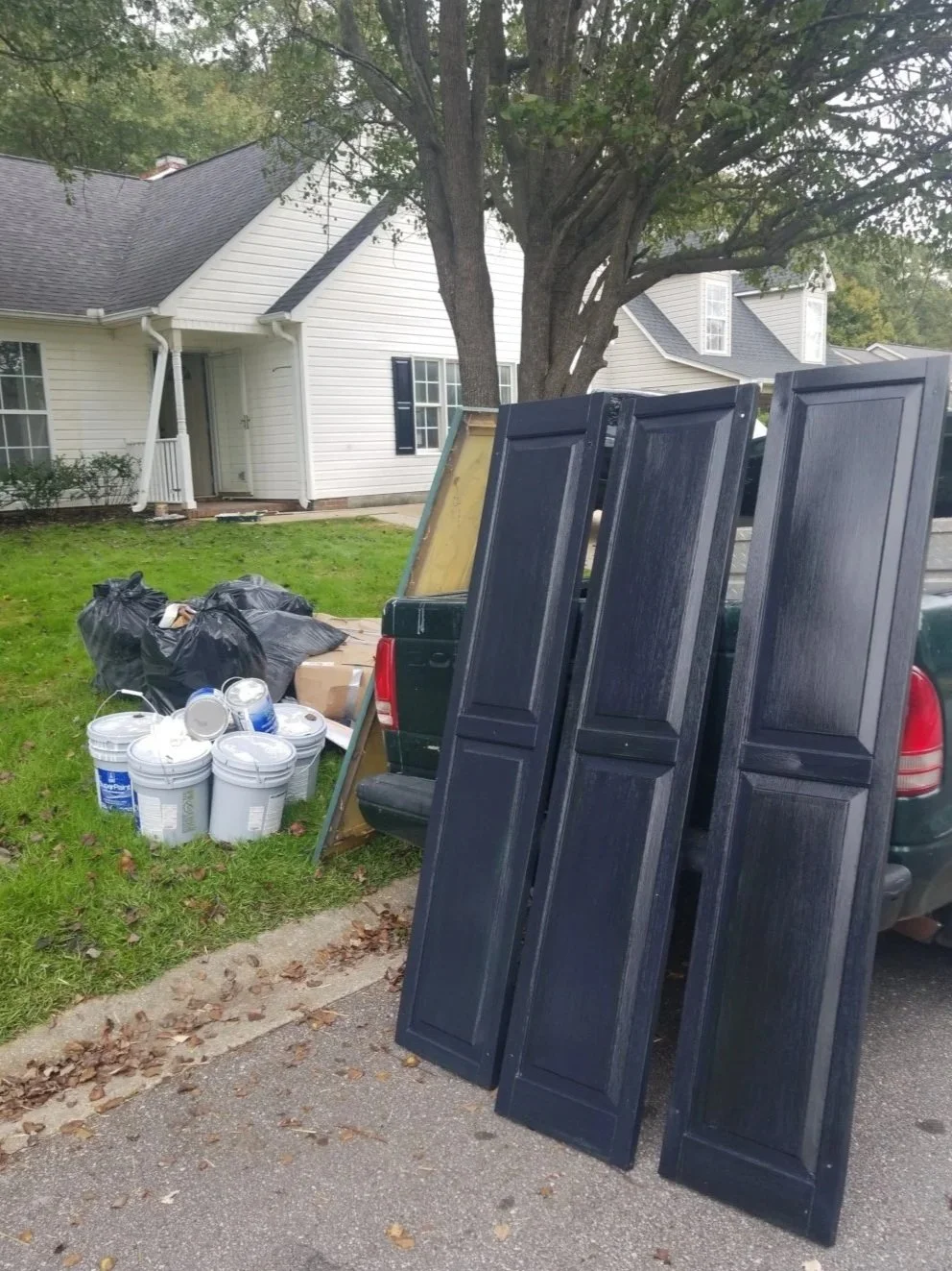 Collection of black cabinet doors leaning against a green truck, with buckets of paint and trash bags nearby, in front of a white house with black shutters and a large tree.