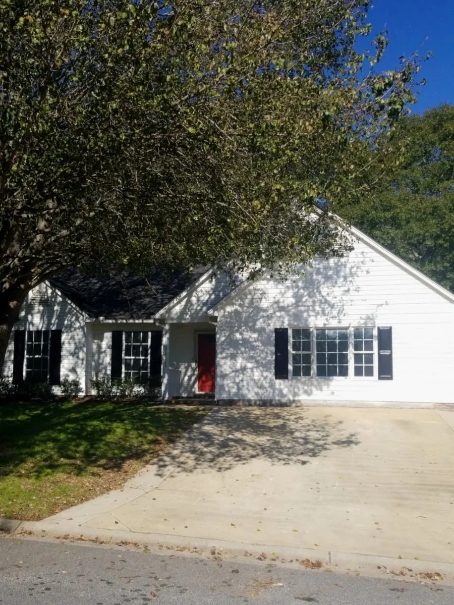 A white house with black shutters, a red front door, large front windows, and a concrete driveway. A large tree with green leaves is casting shadows on the house and driveway. The sky is clear and blue.