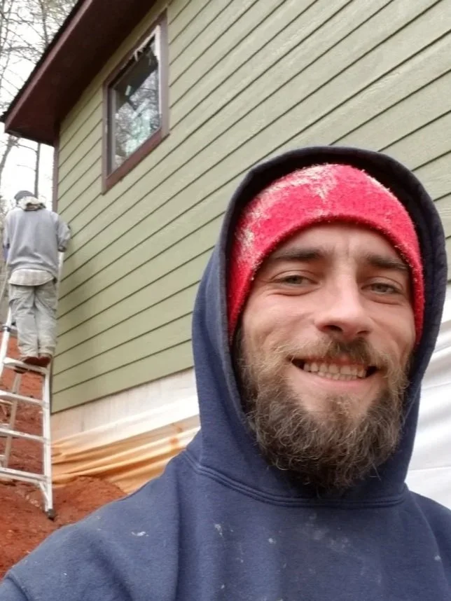 A man taking a selfie near a house under construction, with another worker on a ladder working on the house wall.