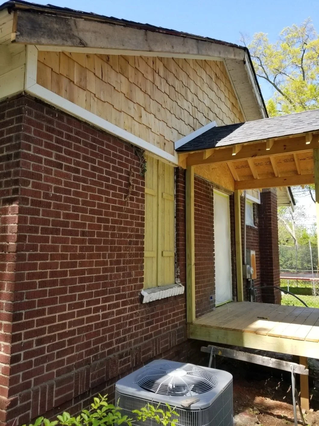 Side view of a house under renovation, showing brick exterior walls, unfinished wooden porch, and new siding project with wood shingles on the upper part. Trees and blue sky are in the background.