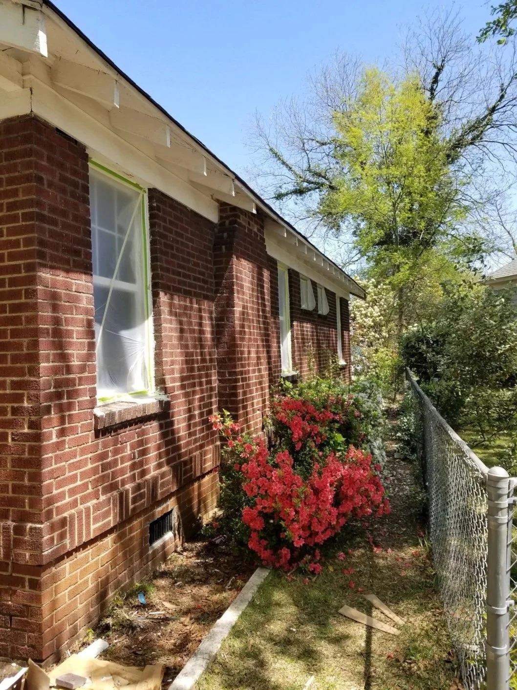 Side view of a red brick house with windows covered in protective plastic. There are blooming pink flowers and a green tree with some leaves just turning green. A chain-link fence runs alongside the house on a sunny day.