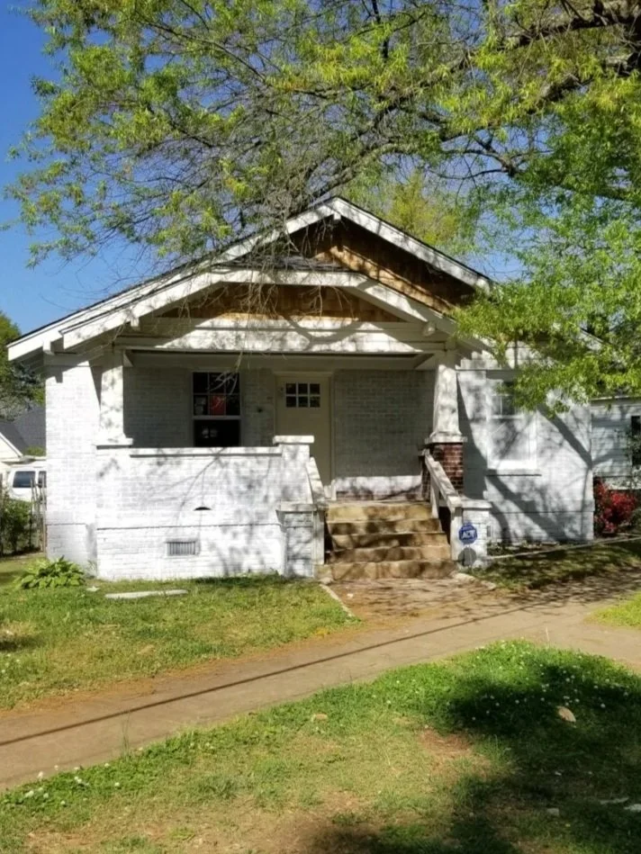 A small white brick house with a front porch, surrounded by green grass and large trees under a blue sky.