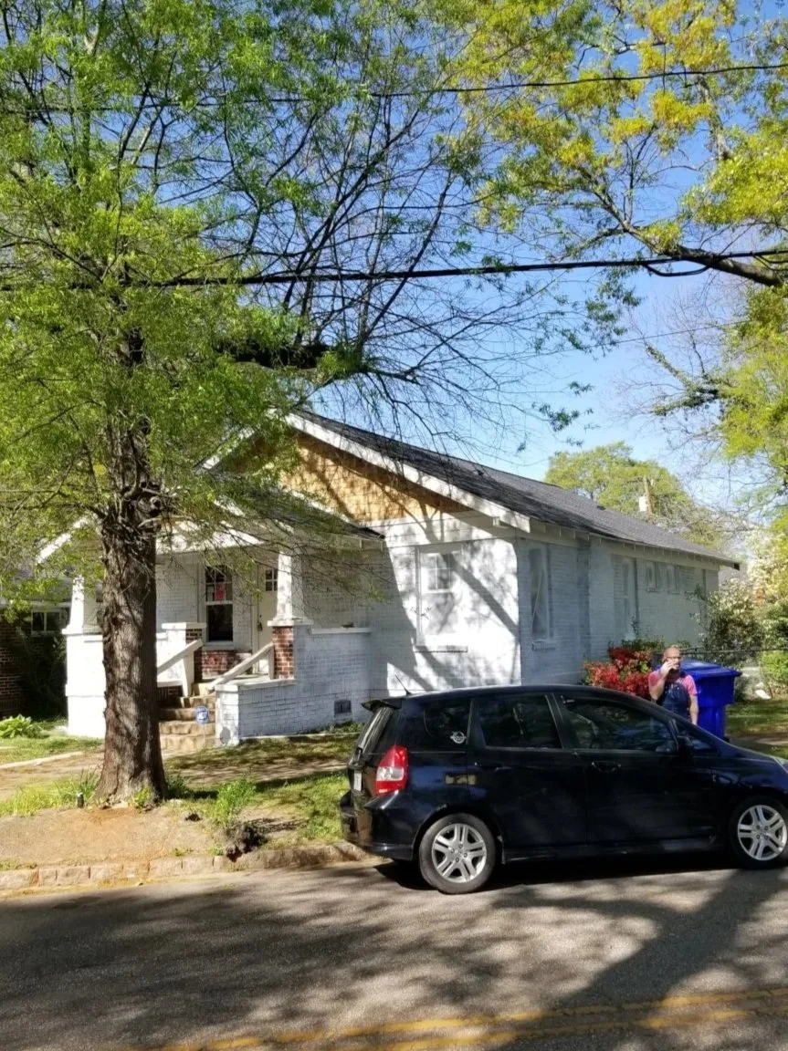 A white house with a porch and stairs, a large tree in front, and a person standing beside a blue trash bin on the sidewalk. A black car is parked on the street.