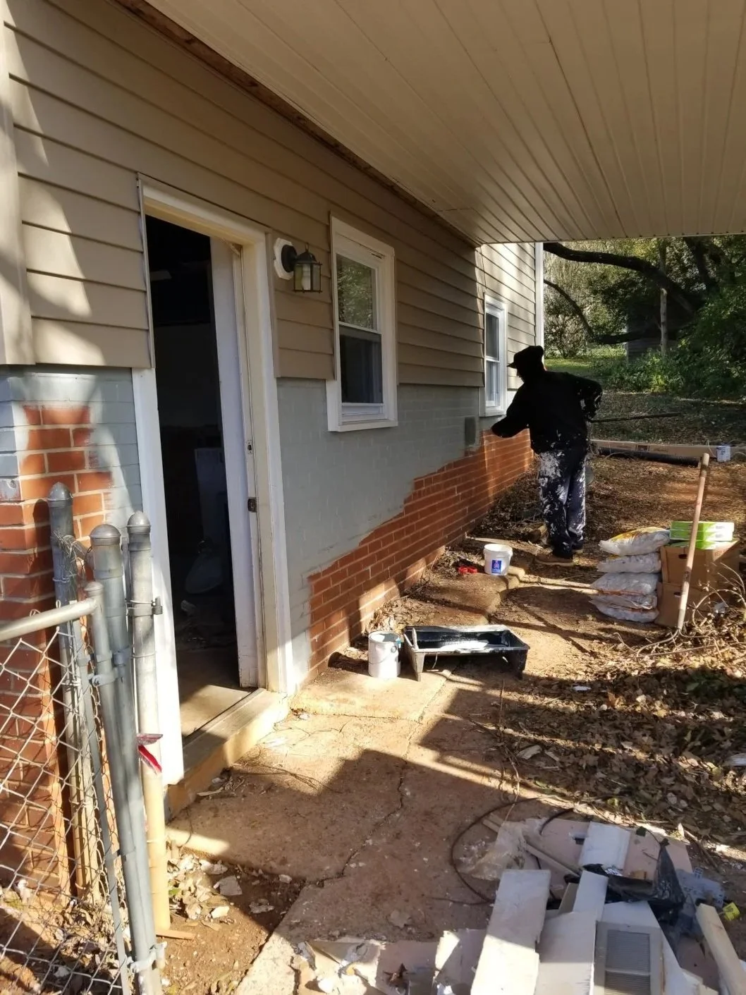 Person painting the exterior wall of a house with beige siding and red bricks, using white paint, in a yard with construction materials and debris.