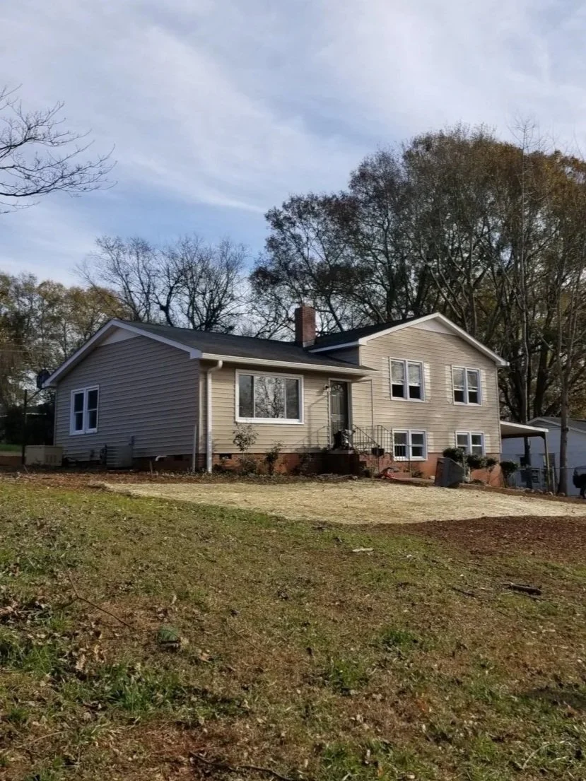 A two-story beige house with white trim, black shutters, and a gable roof, situated on a sloped lawn with trees in the background.