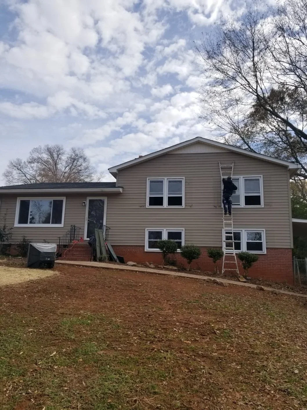 A person ladders up a house to clean or inspect windows on the second floor, with a ladder leaning against the house. The house has beige siding and white-framed windows, with a front yard that has patches of grass and dirt, small bushes, and a cloud