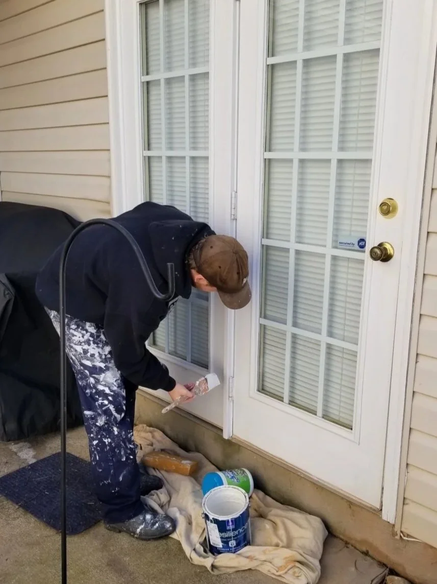 Person painting a white door with a spray brush, dressed in work clothes, outside a house.