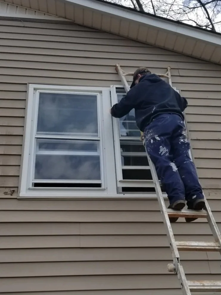 A man in black hoodie and blue work pants standing on a wooden ladder, painting or repairing the exterior window frame of a beige house.