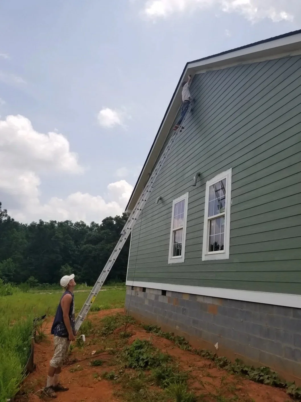 A person standing on the ground holding a ladder, leaning against the side of a house, with another person at the top of the ladder working on the roof.