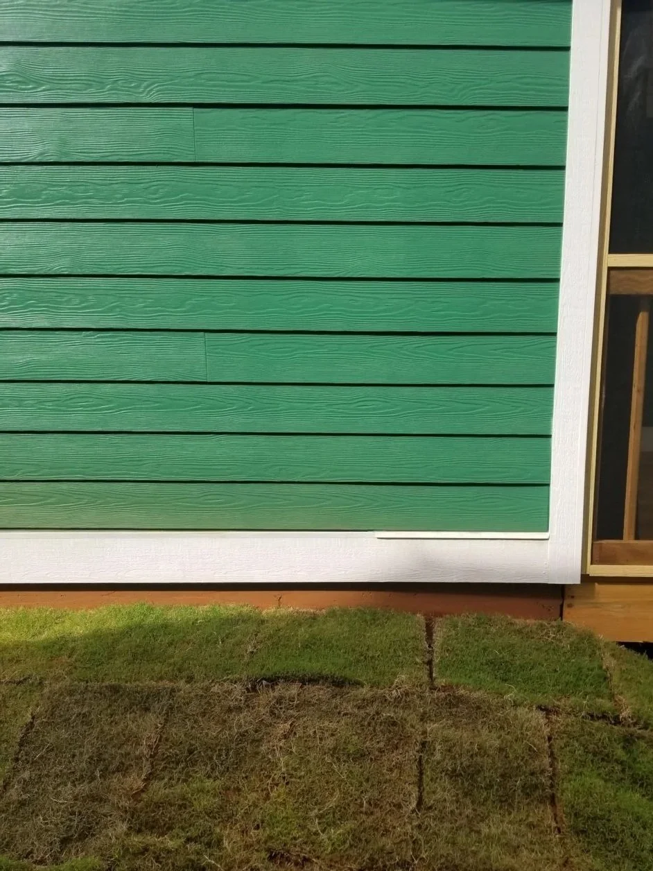 Close-up of green horizontal siding on a building with a white trim at the bottom and a patch of grass in front.