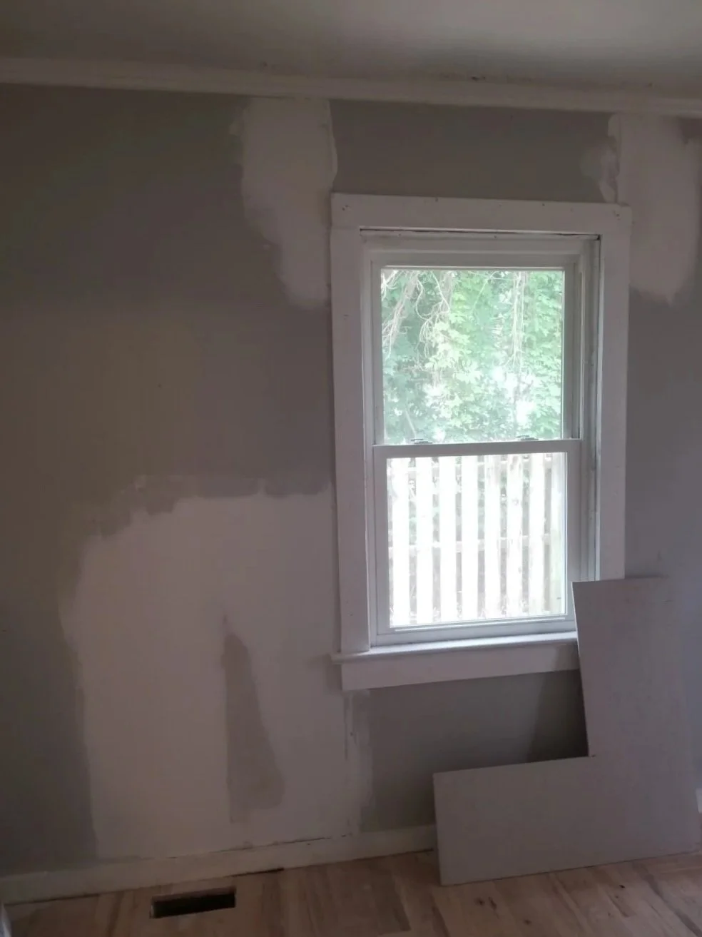 A room under renovation with partially painted or mudded walls, a window with greenery outside, and a piece of drywall leaning against the wall.