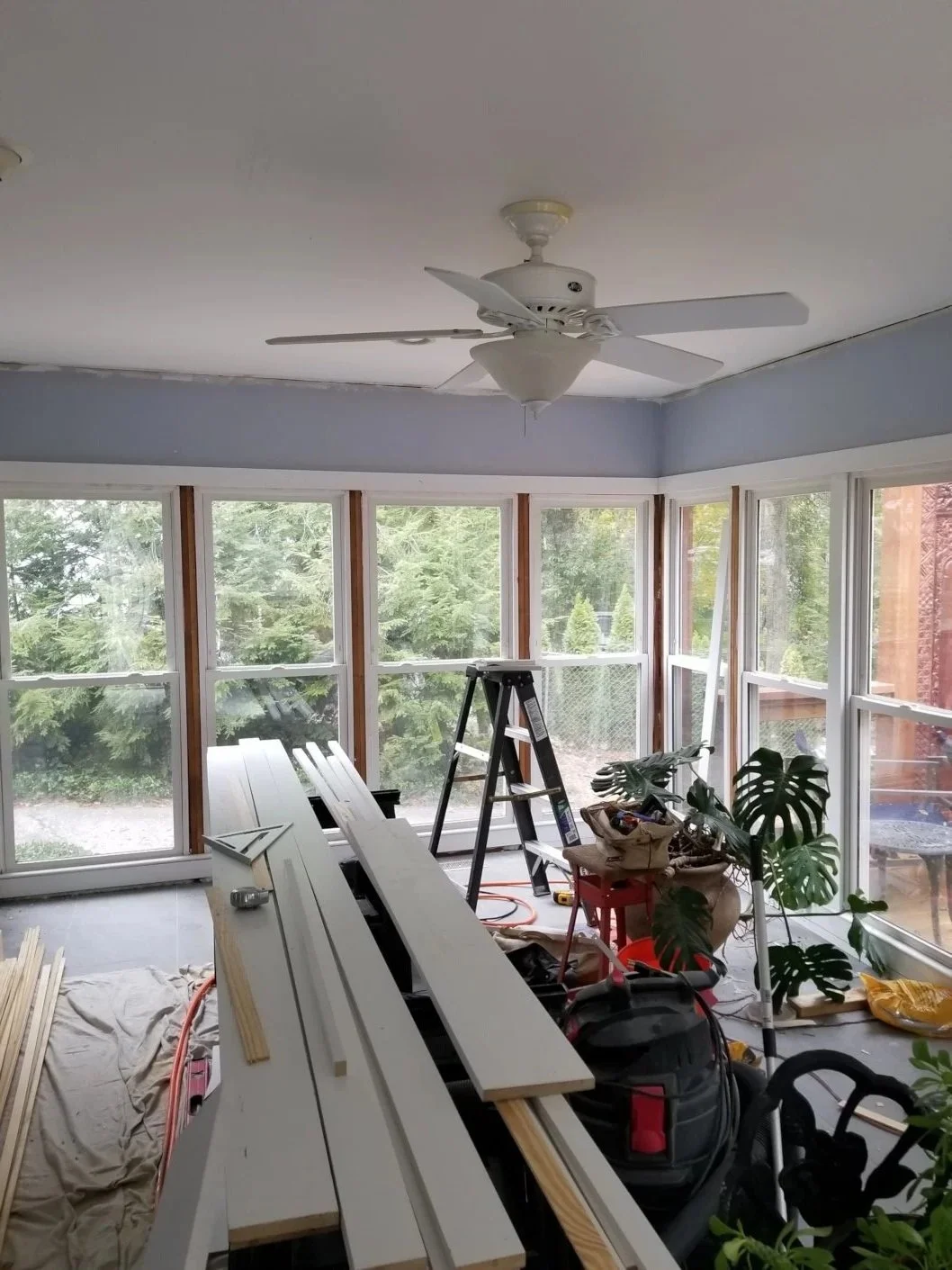 Sunroom under renovation with construction tools, wood planks, a ladder, and potted plants. Large windows reveal green trees outside.