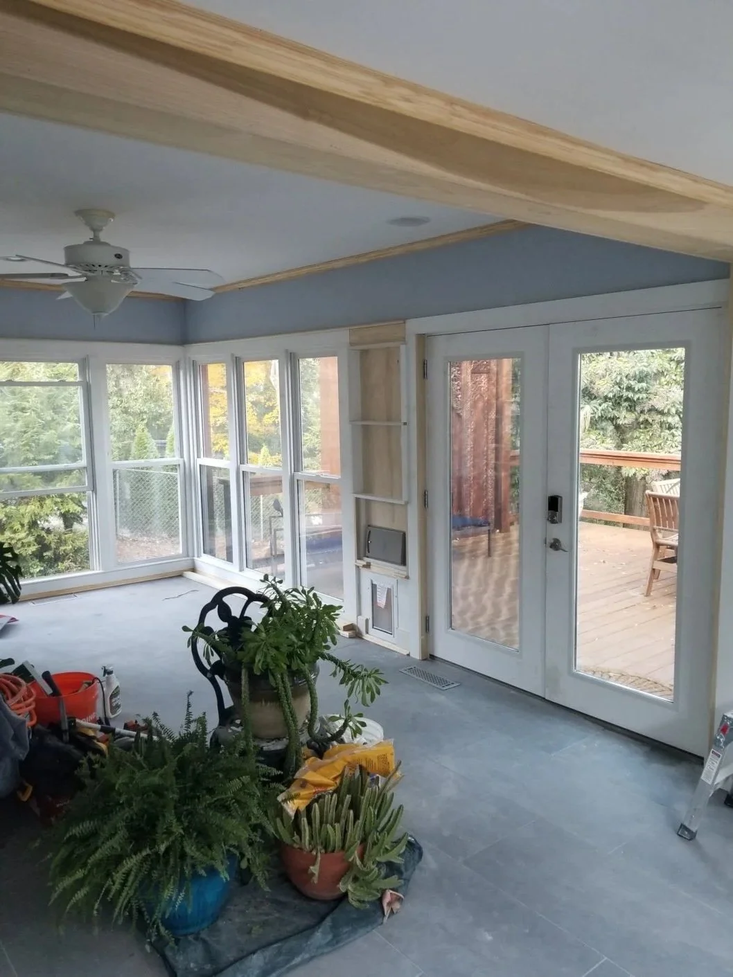 Bright sunroom with large windows and a glass door leading to a wooden deck, ceiling fan, and several potted plants on the floor.