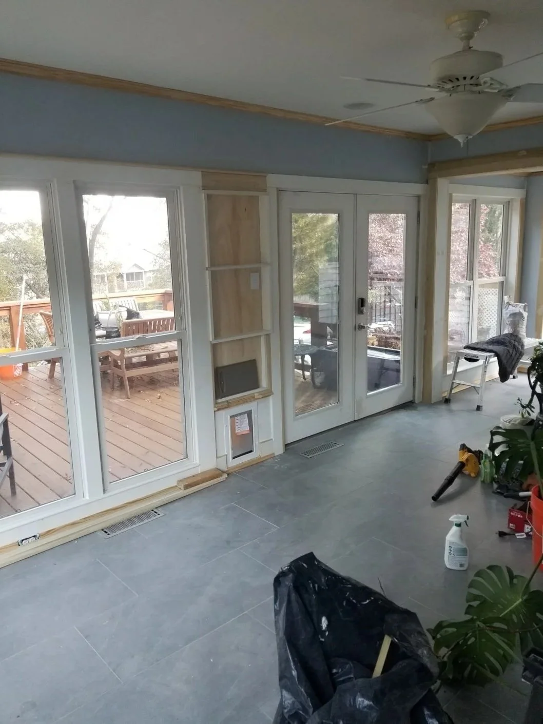 A room with large windows and glass doors leading to a wooden deck outside. The interior appears to be under renovation with tools, a spray bottle, and materials scattered around. There is a ceiling fan and some built-in shelving next to the doors.