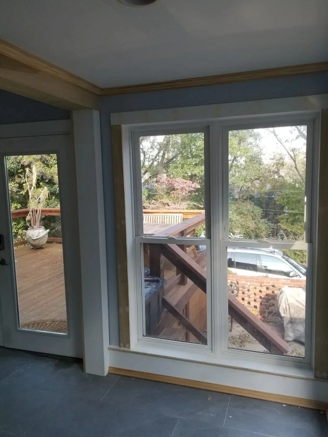 Interior view of a room with a large window and a door leading to a deck outside, showing trees and a parked vehicle.