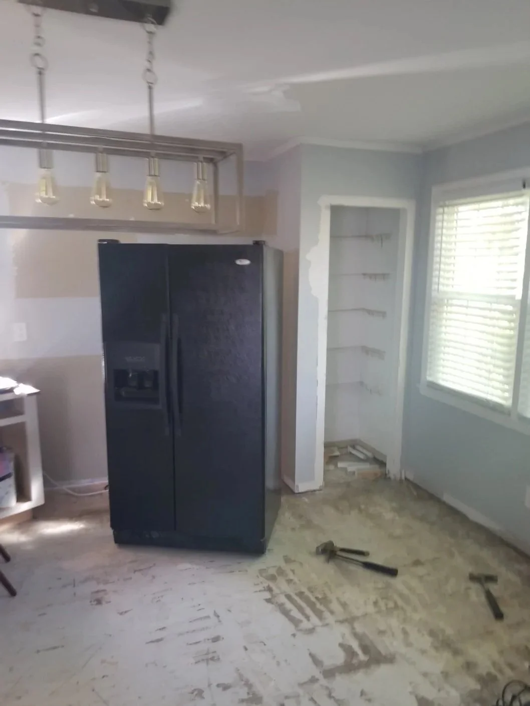 Kitchen with a black refrigerator, unpainted walls, and unfinished flooring. There is an empty built-in shelf space and some tools on the floor.