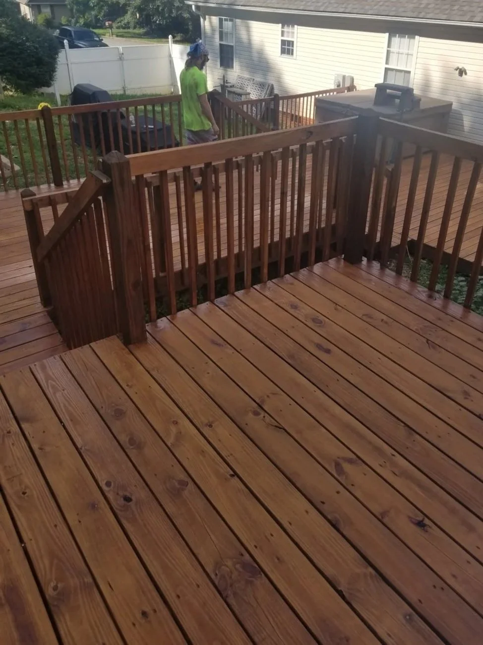 A wooden deck with a railing and stairs, a person wearing a neon green shirt and a blue bandana walking on the deck, in a backyard with a white fence and a house with white siding.