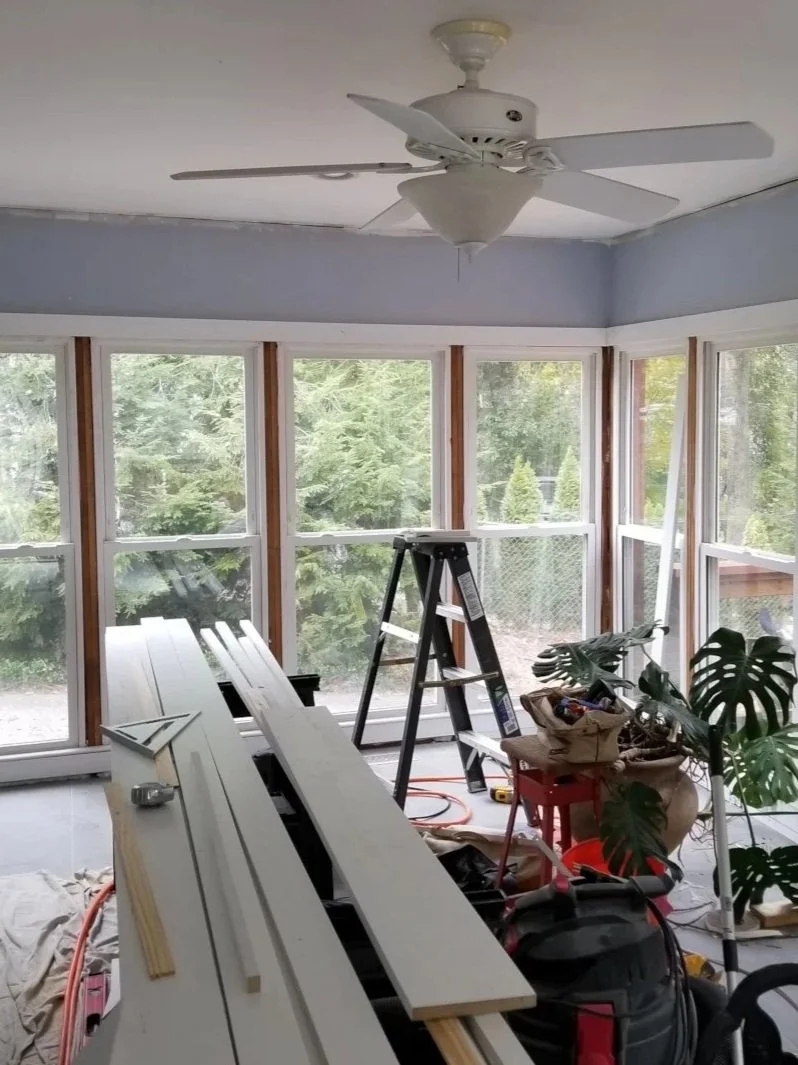 A room under renovation with large windows, a ceiling fan, construction materials, step ladder, and tools.