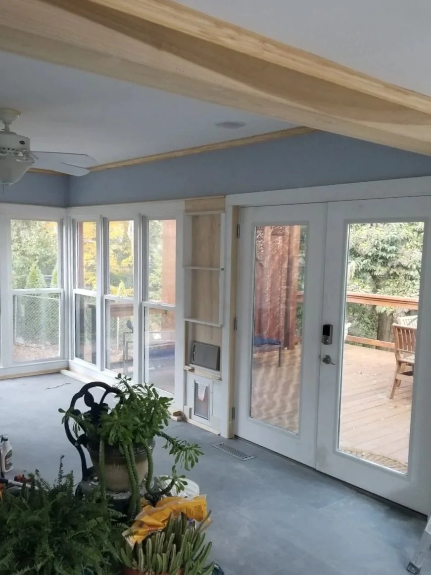 A sunroom with large windows and a glass door leading to a wooden deck outside. The room has a light blue ceiling with a ceiling fan, wooden crown molding, and some houseplants near the door. There are gardening supplies on the floor.