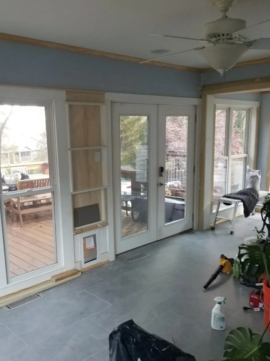Interior of a room under renovation with large glass doors leading to an outdoor deck, a ceiling fan, construction tools, and supplies visible on the floor.