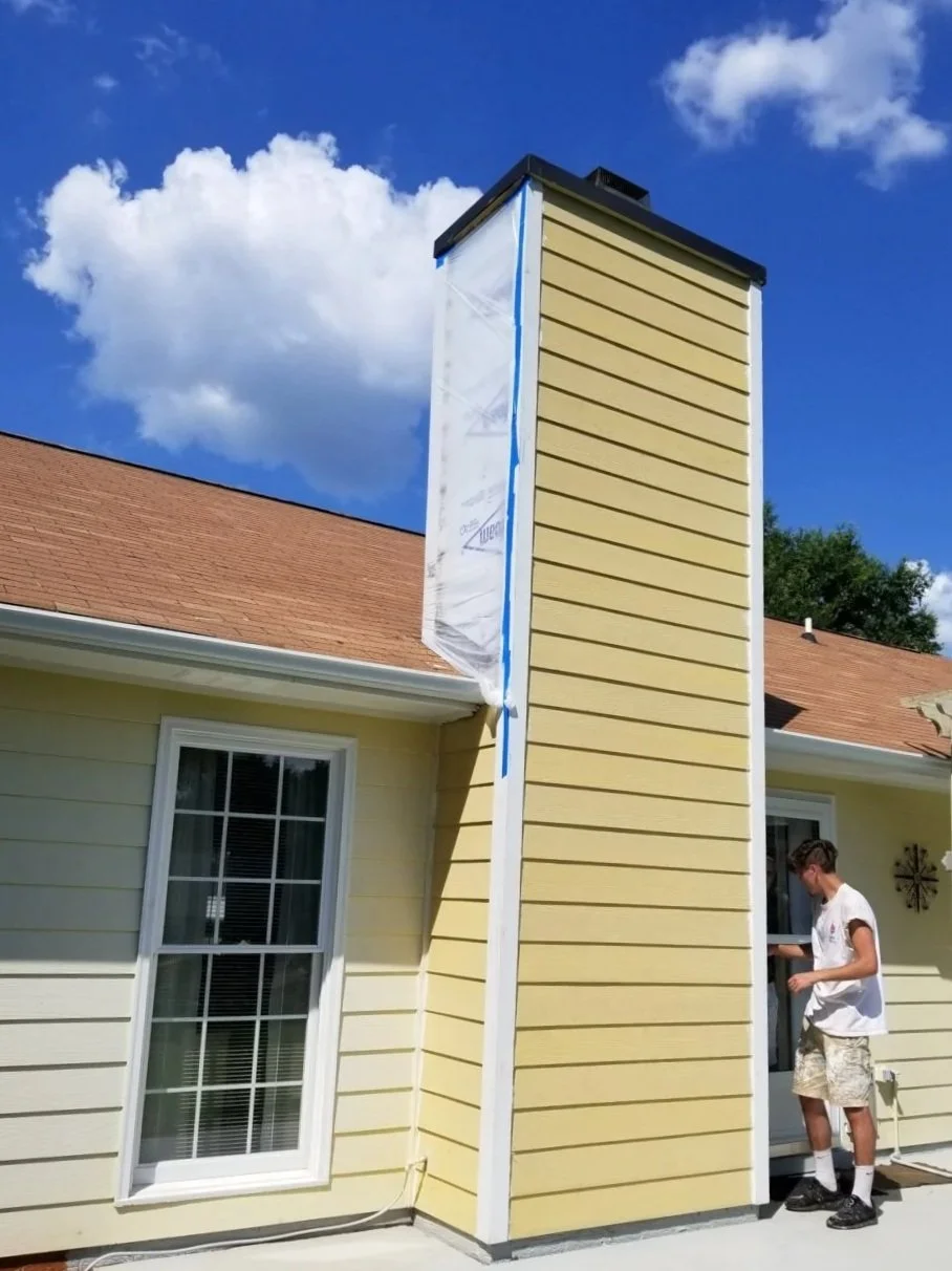 A person working on siding repair or installation on the exterior of a yellow house, near a chimney. The sky is partly cloudy with a bright blue background.