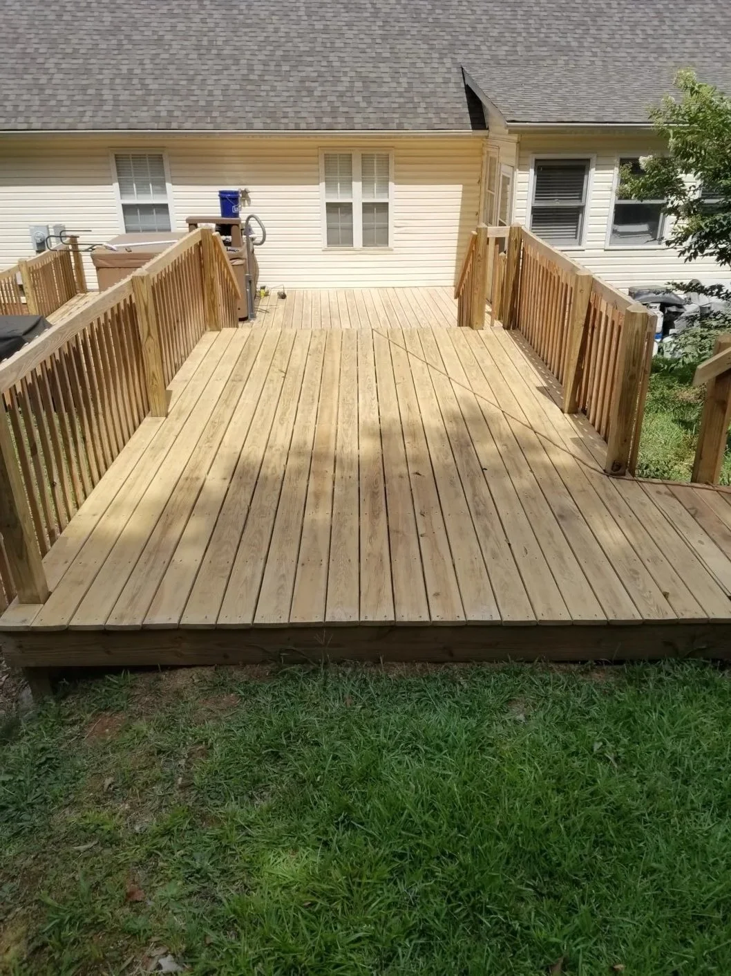 Freshly built wooden deck with railings attached to the back of a house, overlooking a grassy yard with some trees and a neighboring house visible.