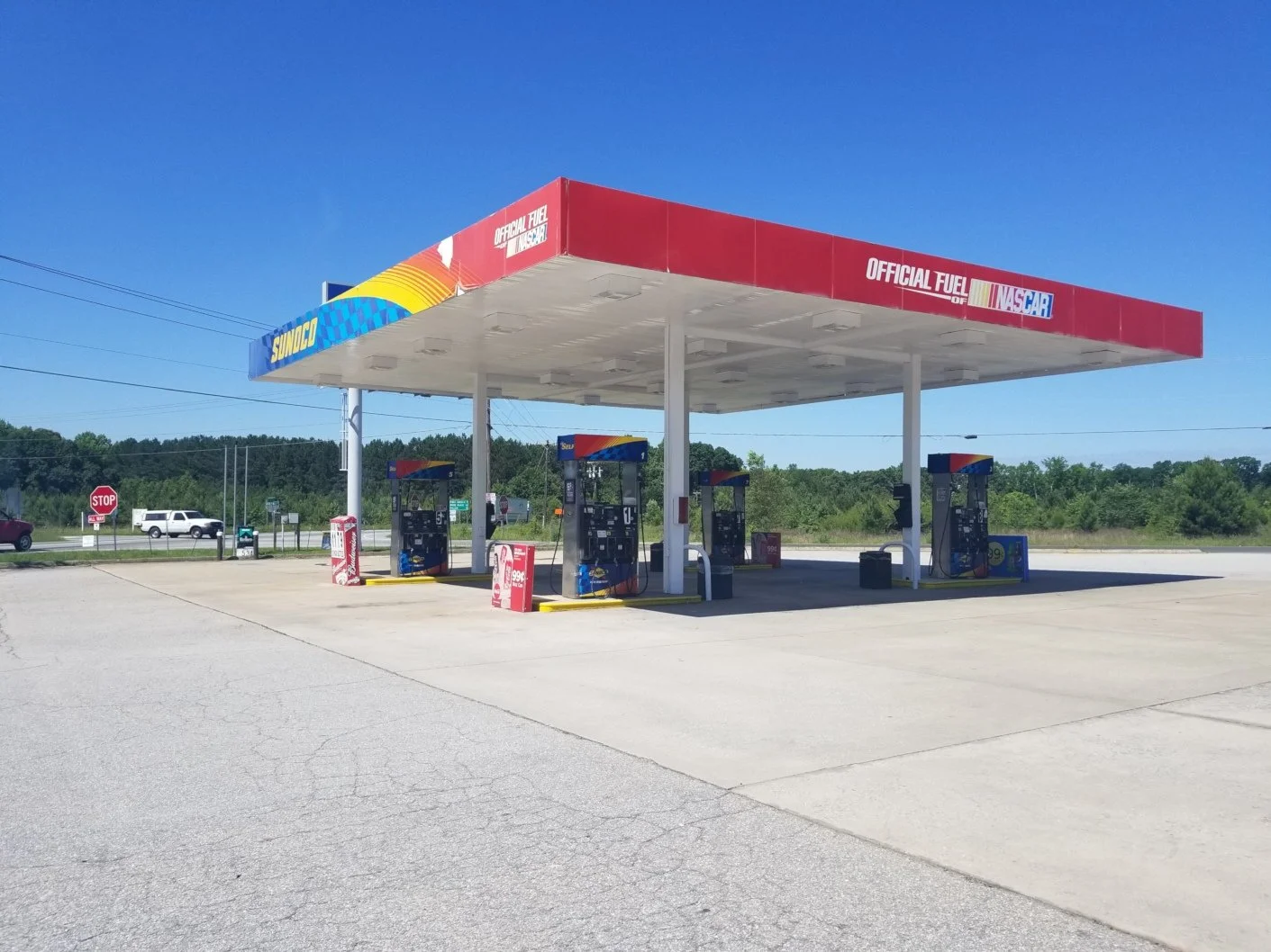 A Sunoco gas station with four fuel pumps under a large red and white canopy, situated in a rural area with a clear blue sky and trees in the background.