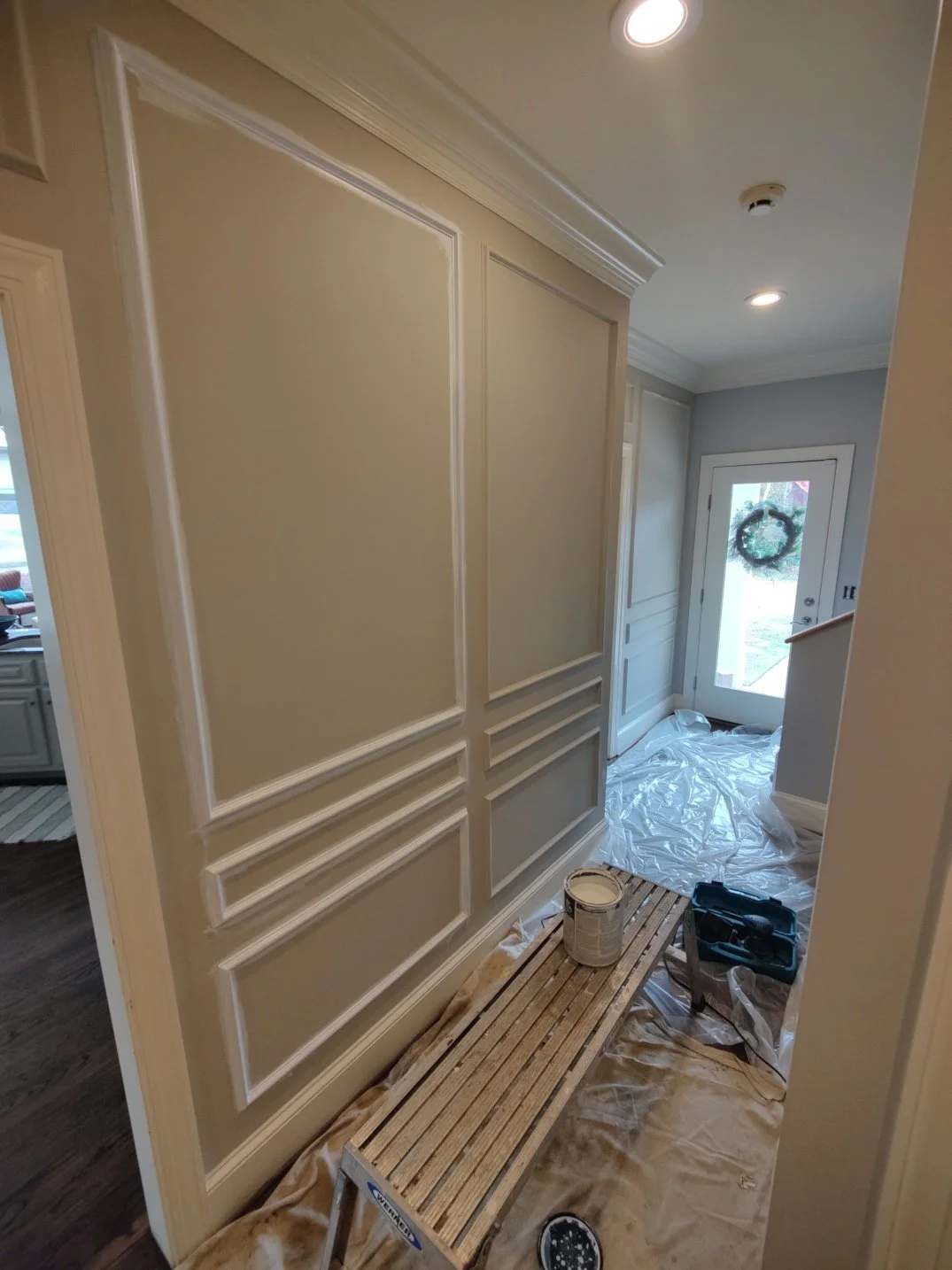 Interior of a home with beige walls and crown molding, a door with a holiday wreath, and painting supplies including a paint can, a toolkit, and a wooden bench on a protective drop cloth.