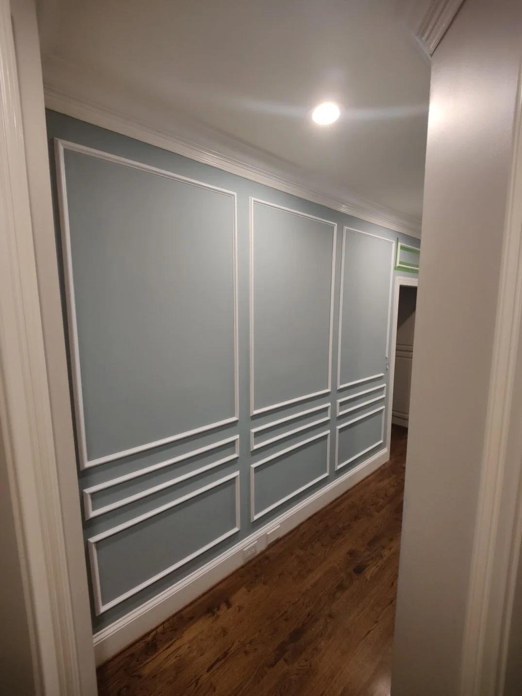Interior hallway with light blue wall panels adorned with white trim and decorative molding, hardwood flooring, and ceiling recessed lighting.