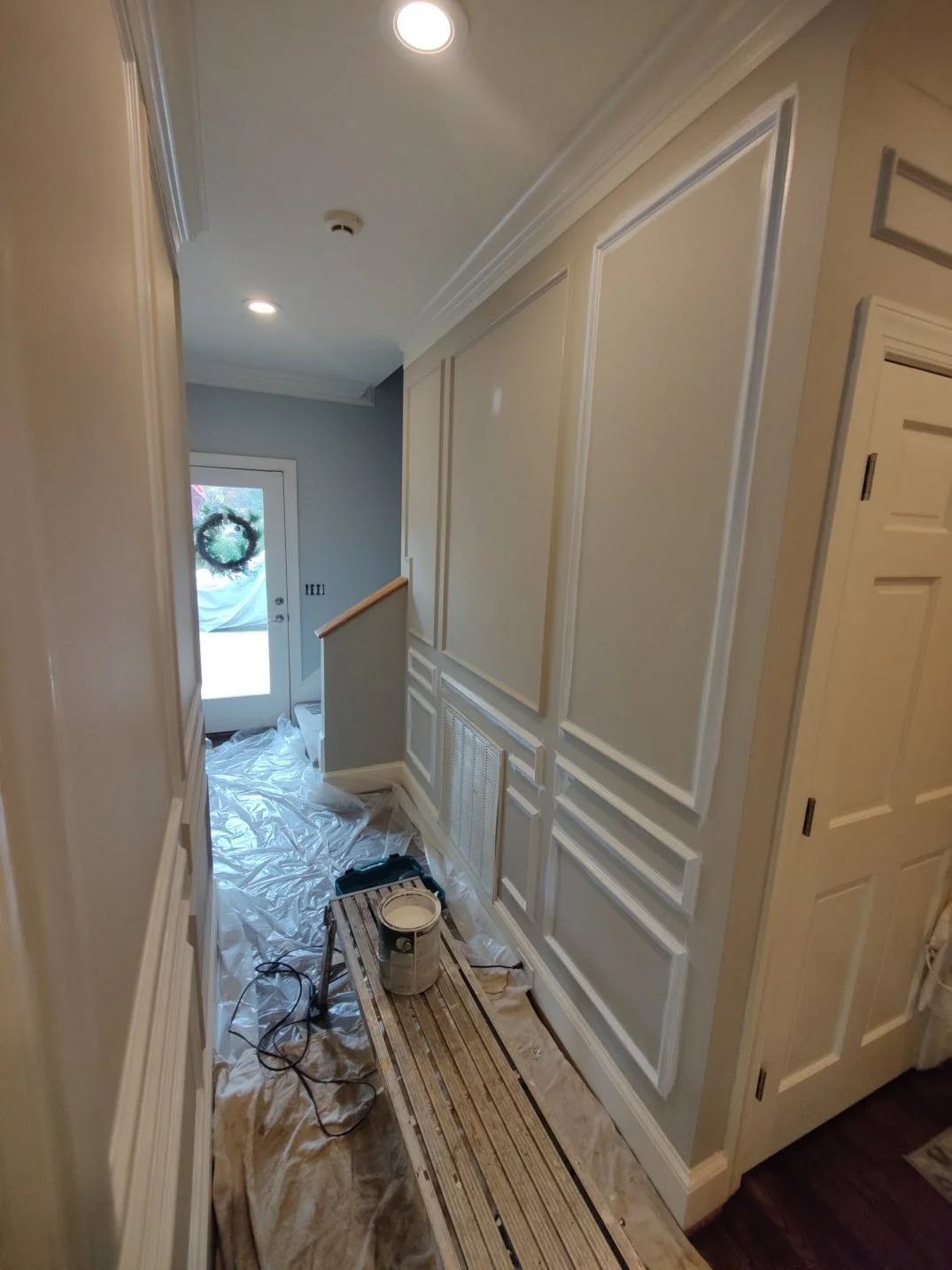 Hallway with beige wainscoting, a staircase leading up, and a door with a wreath at the entrance, with painting tools and plastic sheets on the floor, indicating ongoing painting or renovation.