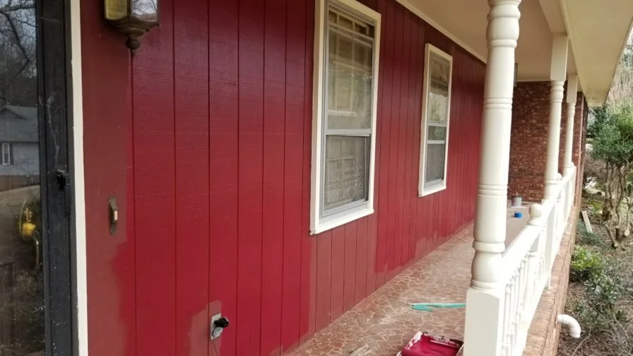 Exterior view of a house with red wooden siding, white-framed windows, a porch with white pillars, and parts of a garden with trees in the background.