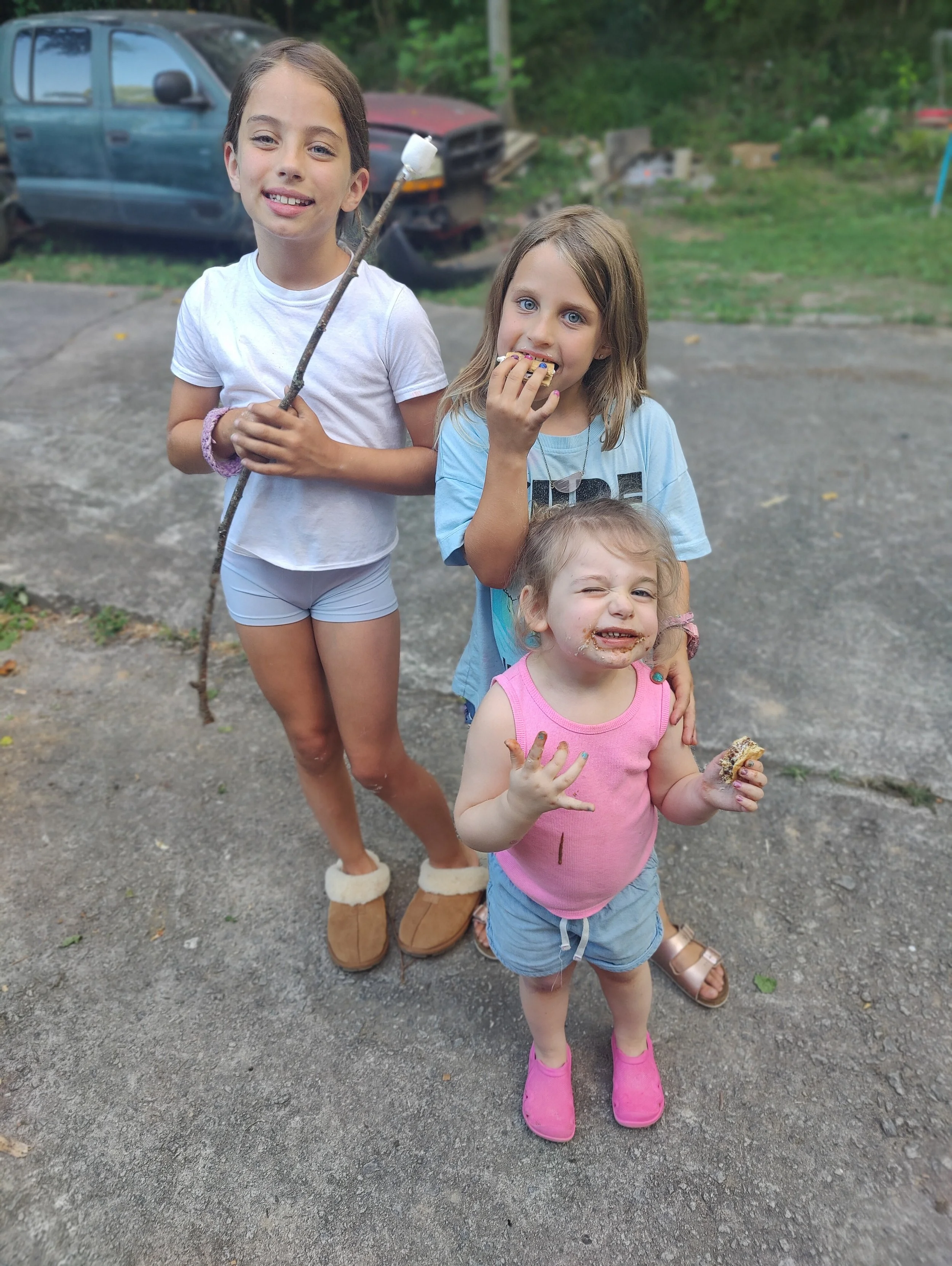 Three young girls standing outside on a driveway, smiling and eating cookies, with a background of cars and greenery.