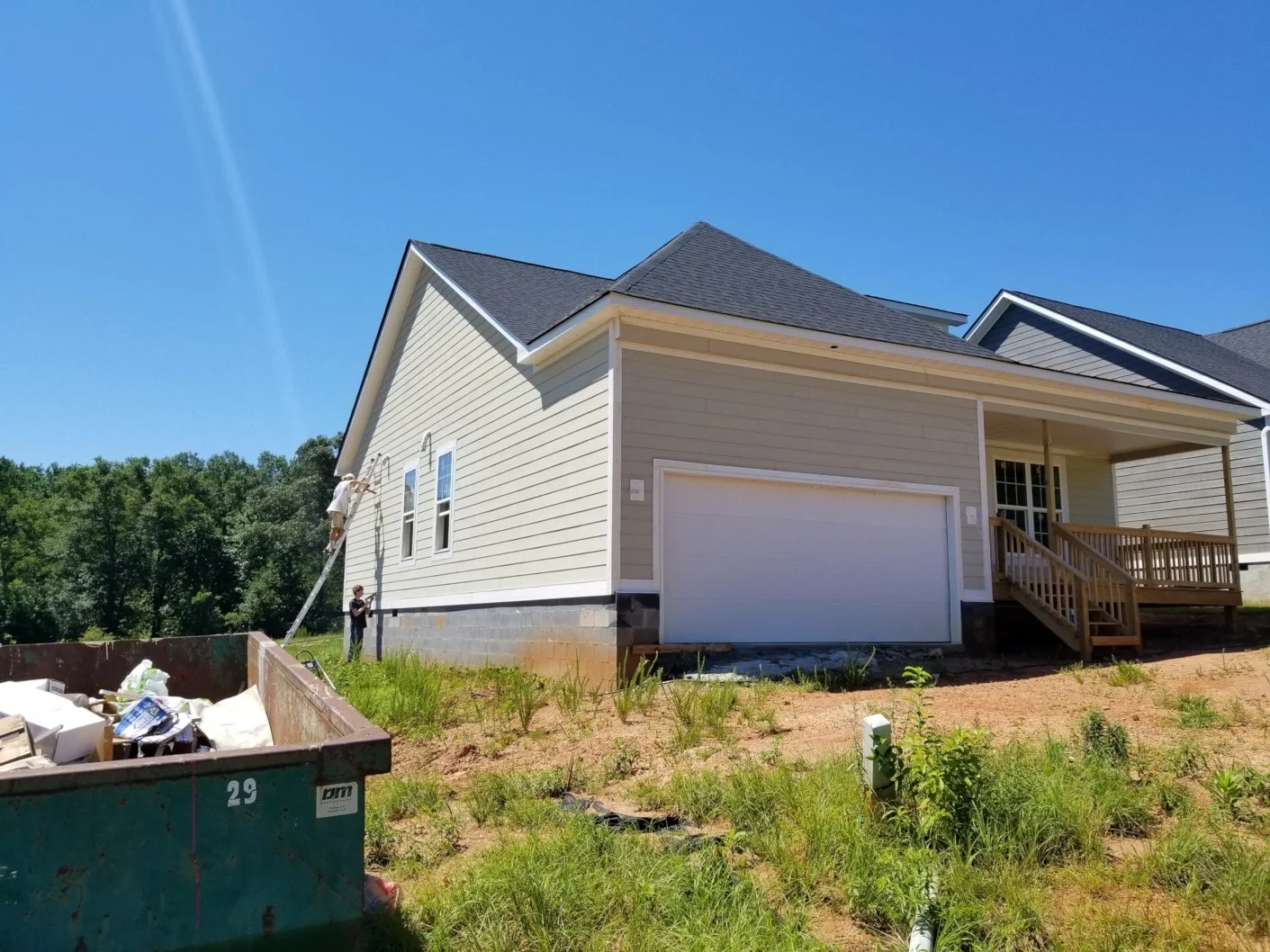 Construction workers painting the exterior wall of a new house on a sunny day, with a yard, a dumpster, and trees in the background.
