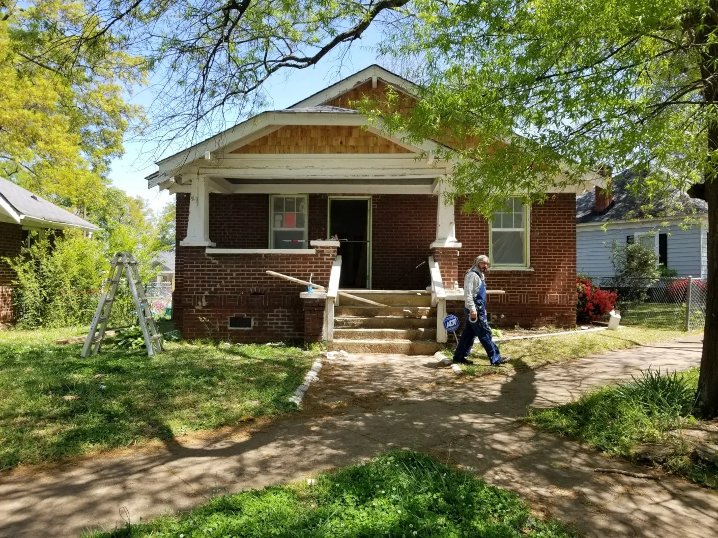 A person wearing a work uniform walking past a house under renovation with stairs missing and construction equipment around, on a sunny day with trees and neighboring houses in the background.