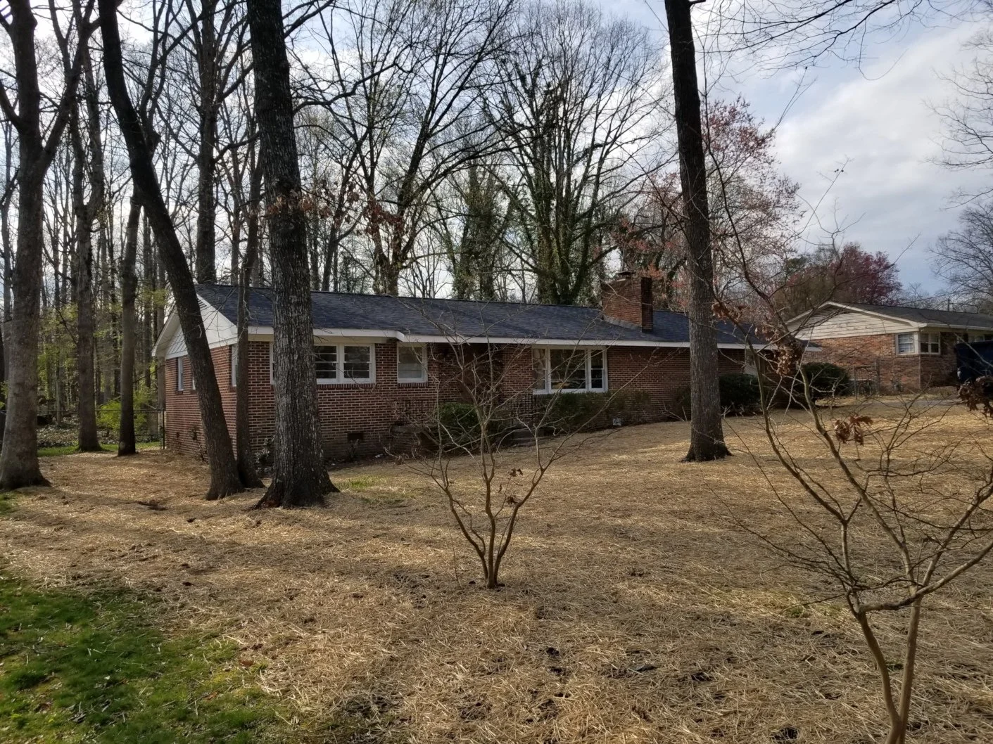 A single-story brick house with a dark roof, surrounded by leafless trees and a brown lawn, on a cloudy day.